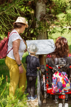 Eine Frau mit Rucksack, ein Kind und eine Frau im Rollstuhl schauen auf eine Ansichtstafel in einem Park.
